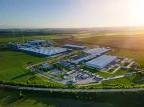 Industry surrounded by green nature, aerial view. Ein großes Fabrikgelände liegt inmitten einer weiten, grünen Landschaft unter einem sonnigen Himmel. Mehrere Gebäude und geparkte Transportfahrzeuge sind sichtbar, umgeben von Feldern und Straßen.