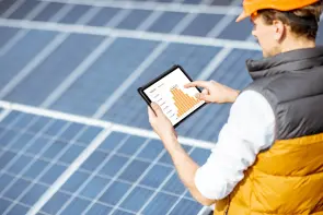 Ein Tablet zeigt ein Balkendiagramm. Ein Mann hält es über Solarpaneln im Freien. Er trägt eine Weste und einen Schutzhelm.
