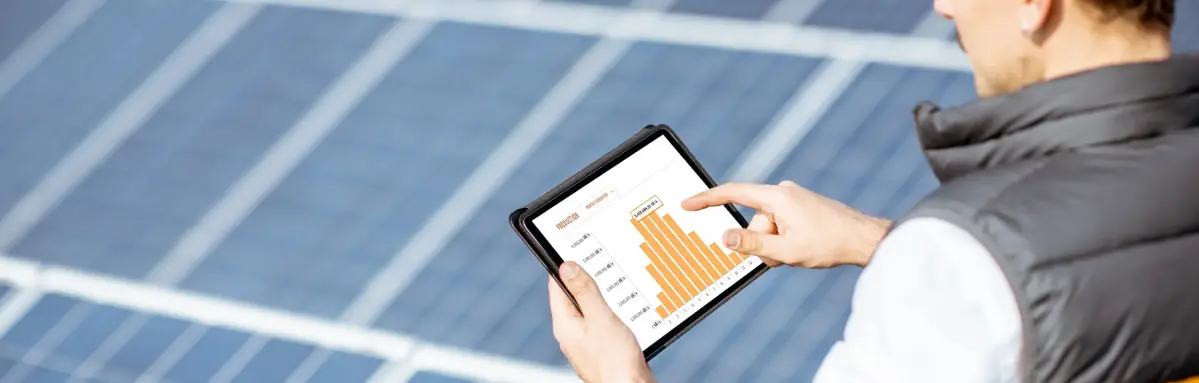 Ein Tablet zeigt ein Balkendiagramm. Ein Mann hält es über Solarpaneln im Freien. Er trägt eine Weste und einen Schutzhelm.