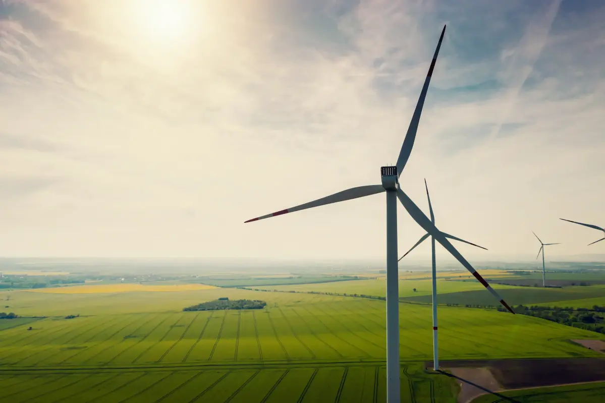 Aerial view of wind turbines and agriculture field Mehrere Windräder drehen sich in einer grünen Landschaft, umgeben von Wäldern und unter einem blauen Himmel mit weißen Wolken.