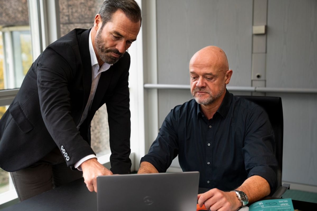 Das Bild zeigt eine Büroszene mit zwei Personen, die gemeinsam an einem Laptop arbeiten. Eine Person sitzt am Tisch vor dem geöffneten Laptop, während die andere Person danebensteht und mit der Hand auf den Bildschirm oder in dessen Richtung zeigt, als würde sie etwas erklären oder besprechen.