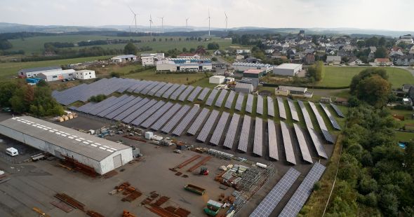 Himmelblick auf einen Solarpark in einer begrünten, bewohnten Umgebung. Daneben eine große Lagerhalle.