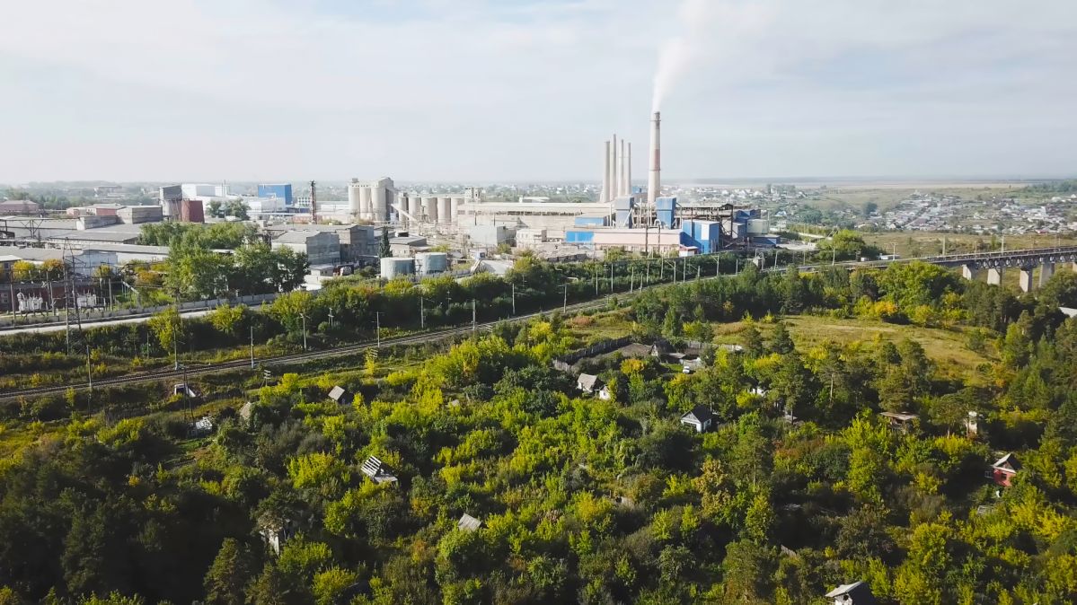 Das Bild zeigt eine große Industrieanlage mit mehreren Schornsteinen, von denen einer Rauch ausstößt. Im Vordergrund befindet sich eine grüne, bewaldete Fläche mit vereinzelten kleinen Gebäuden, dahinter verlaufen Eisenbahngleise. Im Hintergrund erstreckt sich eine Stadtlandschaft unter einem leicht bewölkten Himmel.