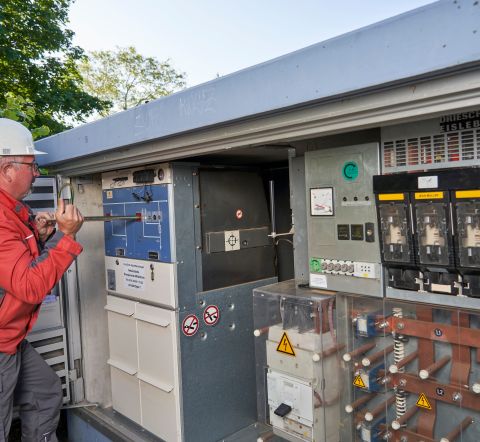 Ein Mann in roter Arbeitskleidung und Schutzhelm arbeitet an einer Transformatorenstation.