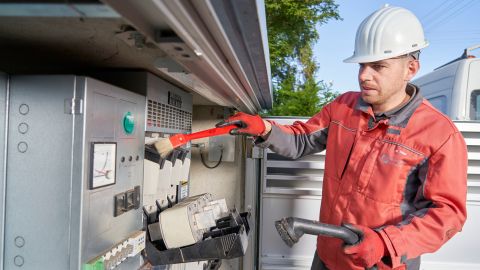 Ein Mann in roter Arbeitskleidung und Schutzhelm arbeitet an einer Transformatorenstation.