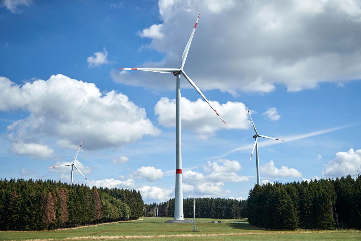 evm-fest im Windpark Höhn zur Erweiterung Drei Windkraftanlagen drehen sich in einer grünen Landschaft, umgeben von Wäldern und unter einem blauen Himmel mit weißen Wolken.