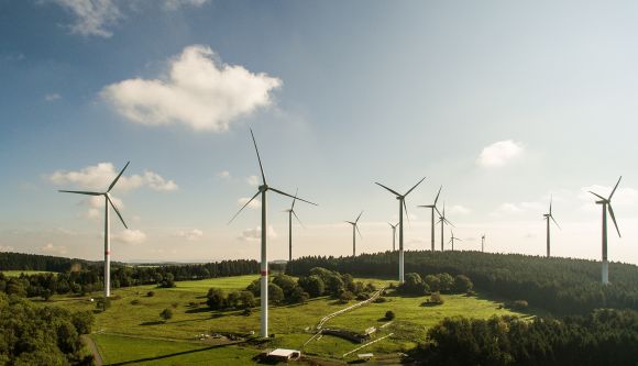 Mehrere Windräder drehen sich in einer grünen Landschaft, umgeben von Wäldern und unter einem blauen Himmel mit weißen Wolken.