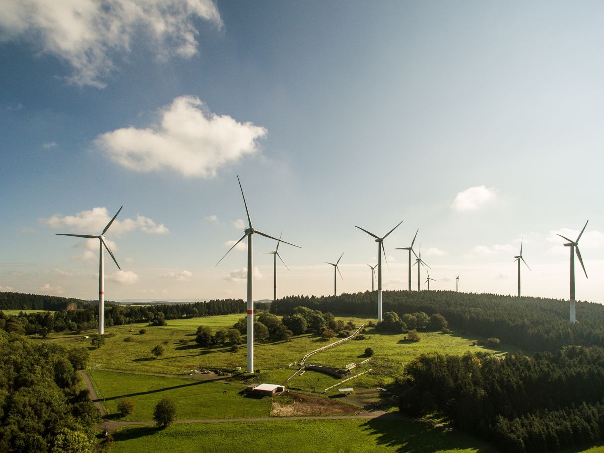 Waigandshain_DJI_0879_opt_A5 Mehrere Windräder drehen sich in einer grünen Landschaft, umgeben von Wäldern und unter einem blauen Himmel mit weißen Wolken.