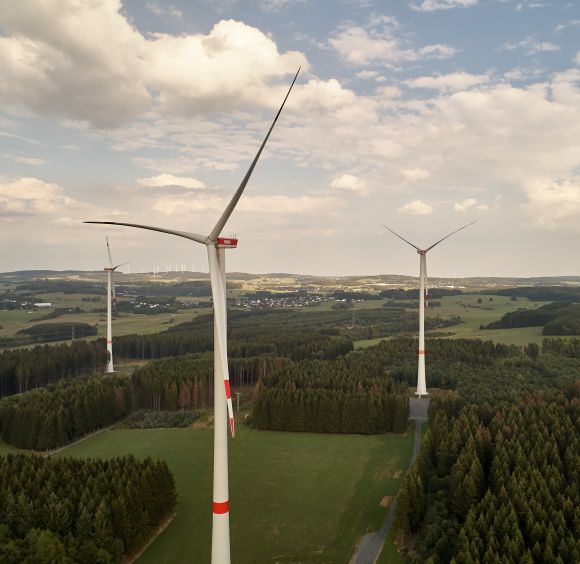 Mehrere Windräder drehen sich in einer grünen Landschaft, umgeben von Wäldern und unter einem blauen Himmel mit weißen Wolken.