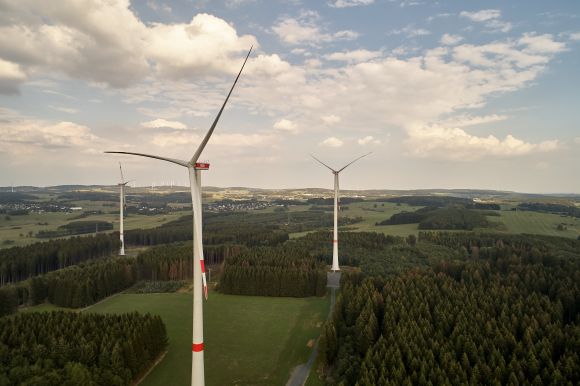 Mehrere Windräder drehen sich in einer grünen Landschaft, umgeben von Wäldern und unter einem blauen Himmel mit weißen Wolken.