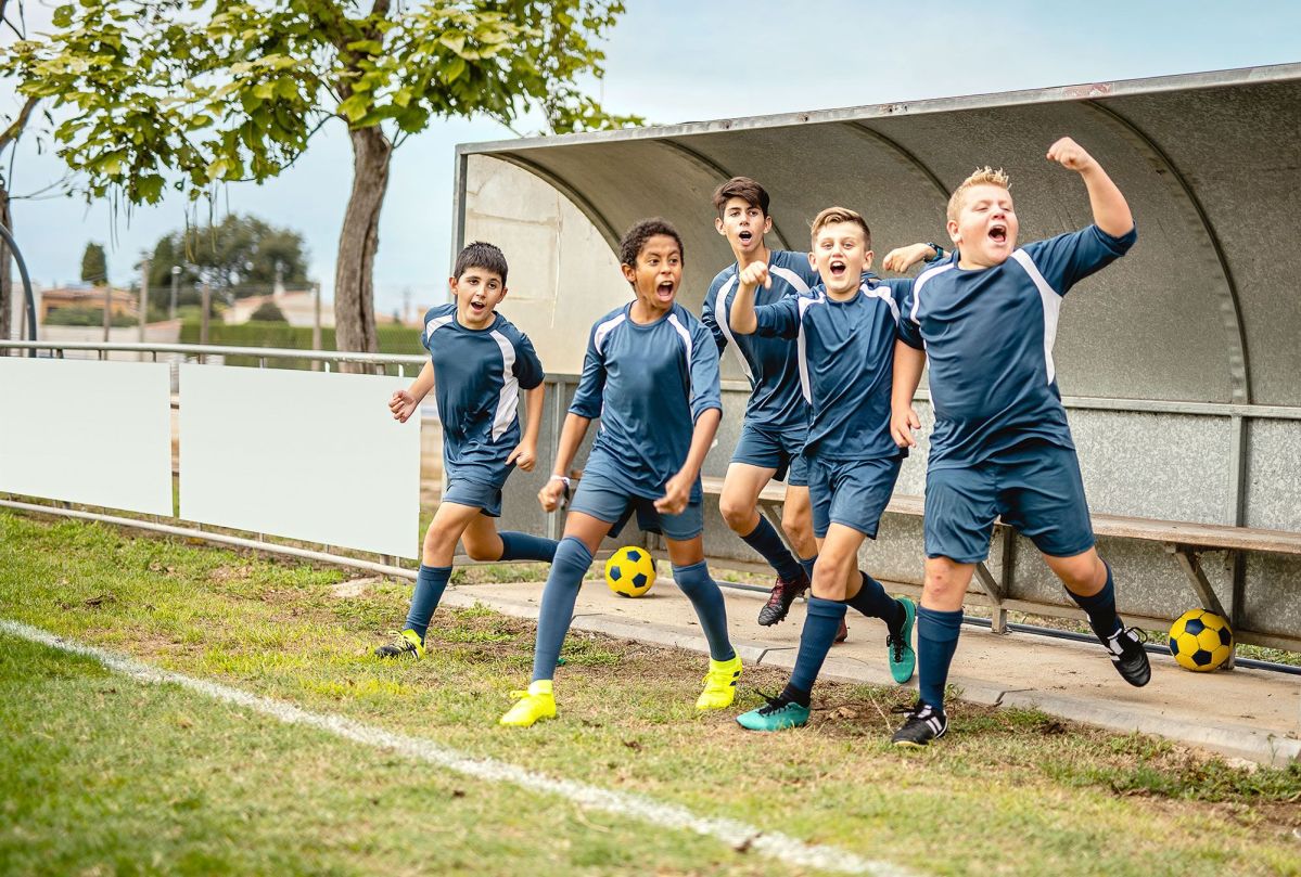 Fünf Jungs in dunkelblauen Fußballuniformen springen fröhlich von einer Bank auf einem Fußballplatz.