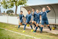 Fünf Jungs in dunkelblauen Fußballuniformen springen fröhlich von einer Bank auf einem Fußballplatz.