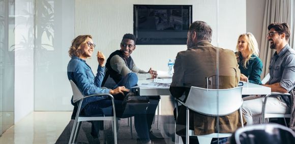 Konferenz im Büro Eine Gruppe von fünf Personen sitzt um einen Tisch und spricht miteinander in einem modernen, hellen Büro mit Glastüren und einem flachen Bildschirm an der Wand.