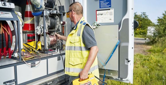 Ein Mann in Warnweste arbeitet an einem offenen Fahrzeug, das mit Kabeltrommeln und Werkzeugen ausgestattet ist. Im Hintergrund befindet sich eine Straße mit Bäumen und Grünflächen.