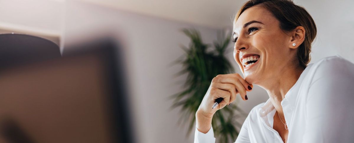 Frau im Büro lacht Eine Frau lächelt entspannt mit einem Stift in der Hand in einem modernen Büro, im Hintergrund ist eine grüne Pflanze verschwommen zu sehen.