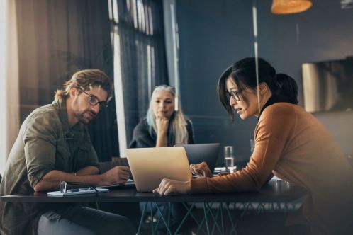 Drei Menschen sitzen an einem Tisch, arbeiten konzentriert an Laptops. Im Hintergrund sind Vorhänge und ein Fernseher sichtbar. Ein Glas Wasser steht auf dem Tisch.
