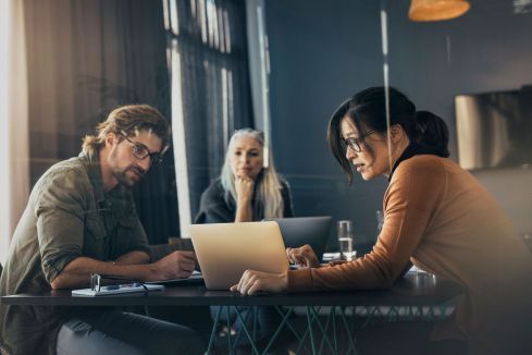 Drei Menschen sitzen an einem Tisch, arbeiten konzentriert an Laptops. Im Hintergrund sind Vorhänge und ein Fernseher sichtbar. Ein Glas Wasser steht auf dem Tisch.