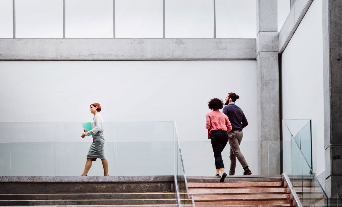 Personen gehen die Treppe hoch Eine Frau geht mit einem Ordner in der Hand an einem Glasgeländer, zwei Personen steigen daneben eine Treppe hinauf in einem modernen, strukturierten Gebäude mit großen Fenstern und Betonwänden.