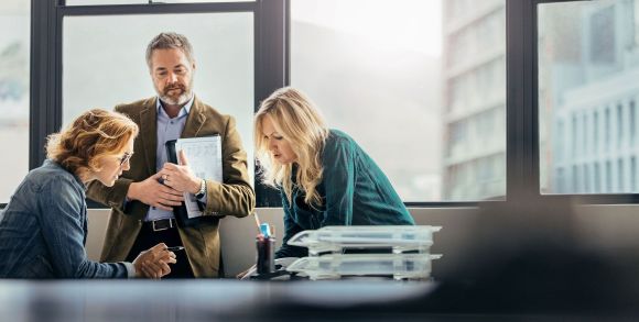 Unterhaltung zwischen Personen Drei Personen besprechen sich in einem Büro, wobei eine Frau sitzt und Notizen macht, während die anderen stehen. Große Fenster im Hintergrund bieten Ausblick auf Gebäude.