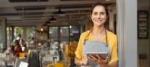 Eine Frau mit einer grauen Schürze steht in einem Café und lächelt freundlich. Sie hält ein Tablet in der Hand.