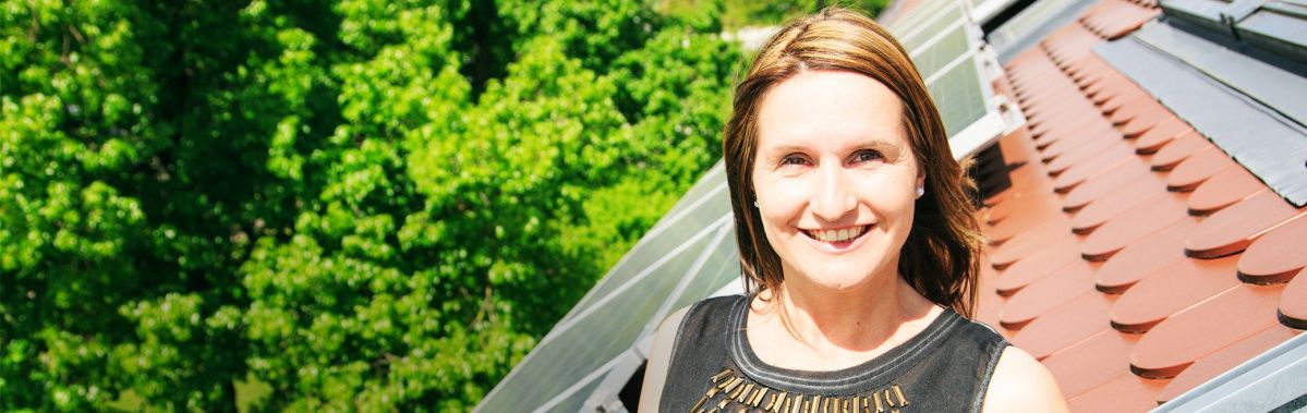 Eine lächelnde Frau steht draußen neben Solarpaneelen. Im Hintergrund ist üppiges grünes Laub zu sehen, das einen sonnigen Tag andeutet.