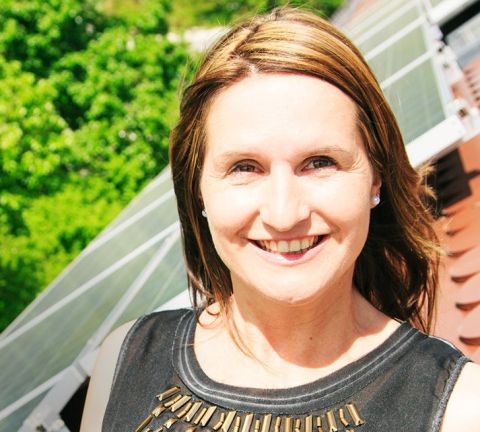 Eine lächelnde Frau steht draußen neben Solarpaneelen. Im Hintergrund ist üppiges grünes Laub zu sehen, das einen sonnigen Tag andeutet.