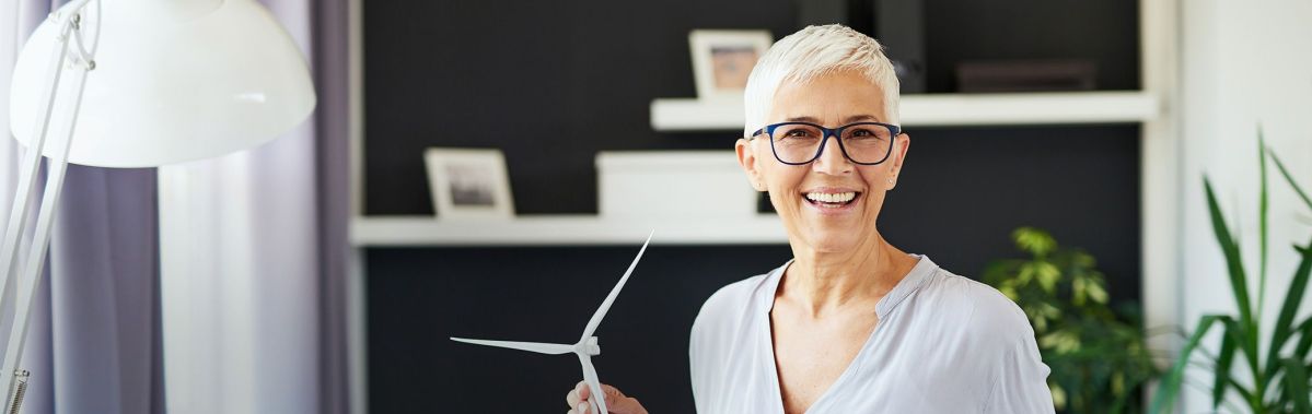 Eine Frau mit kurzen weißen Haaren und Brille hält ein Modell einer Windturbine in der Hand und lächelt in einem modern eingerichteten Raum mit Regalen im Hintergrund.