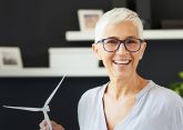 Eine Frau mit kurzen weißen Haaren und Brille hält ein Modell einer Windturbine in der Hand und lächelt in einem modern eingerichteten Raum mit Regalen im Hintergrund.