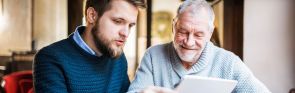 Ein älterer Mann und ein jüngerer Mann betrachten gemeinsam ein Tablet. Sie sitzen drinnen in einem gut beleuchteten Raum, beide wirken konzentriert und interessiert.