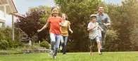 Eine Familie mit zwei kleinen Kindern läuft lachend durch einen Garten. Im Hintergrund steht ein modernes Haus.