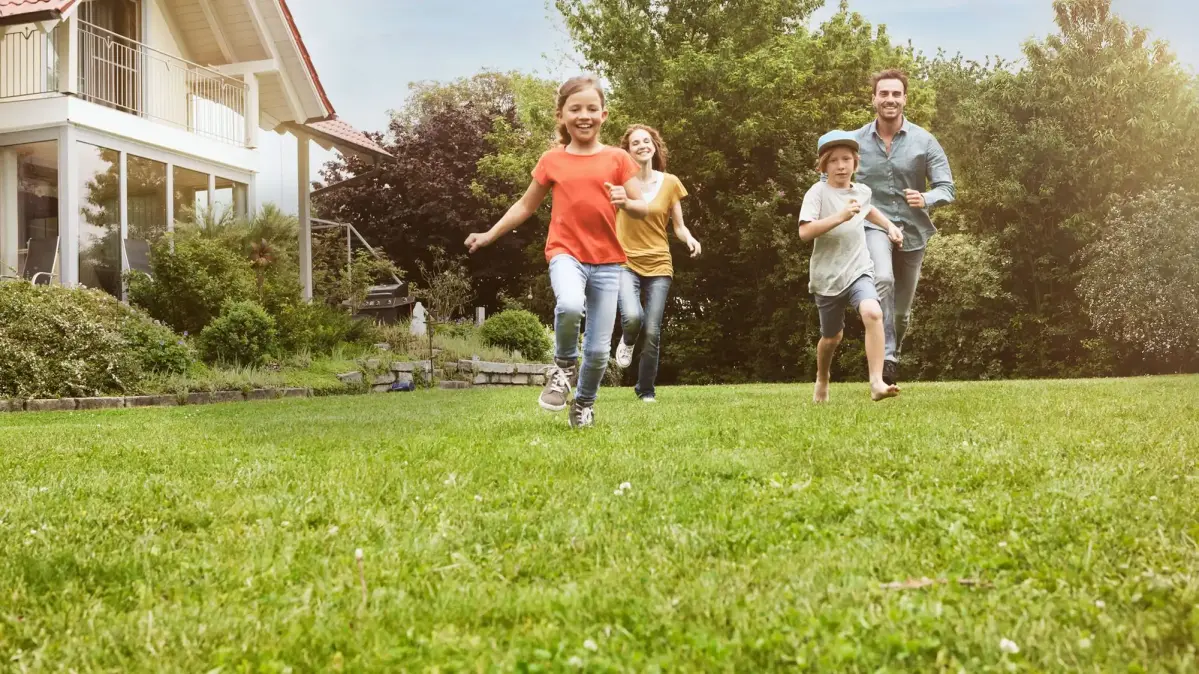 Eine Familie mit zwei kleinen Kindern läuft lachend durch einen Garten. Im Hintergrund steht ein modernes Haus.