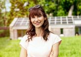 Eine Person lächelt in einer grünen Wiese, trägt eine Sonnenbrille auf dem Kopf. Im Hintergrund stehen Gebäude mit Solarpaneelen, umgeben von Bäumen.