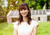 Eine Person lächelt in einer grünen Wiese, trägt eine Sonnenbrille auf dem Kopf. Im Hintergrund stehen Gebäude mit Solarpaneelen, umgeben von Bäumen.