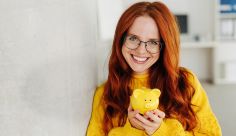 Eine Frau mit langen roten Haaren und Brille hält lächelnd ein gelbes Sparschwein in den Händen. Sie trägt einen gelben Pullover. Der Hintergrund ist ein unscharfes Büro.