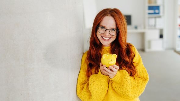 Eine Frau mit langen roten Haaren und Brille hält lächelnd ein gelbes Sparschwein in den Händen. Sie trägt einen gelben Pullover. Der Hintergrund ist ein unscharfes Büro.