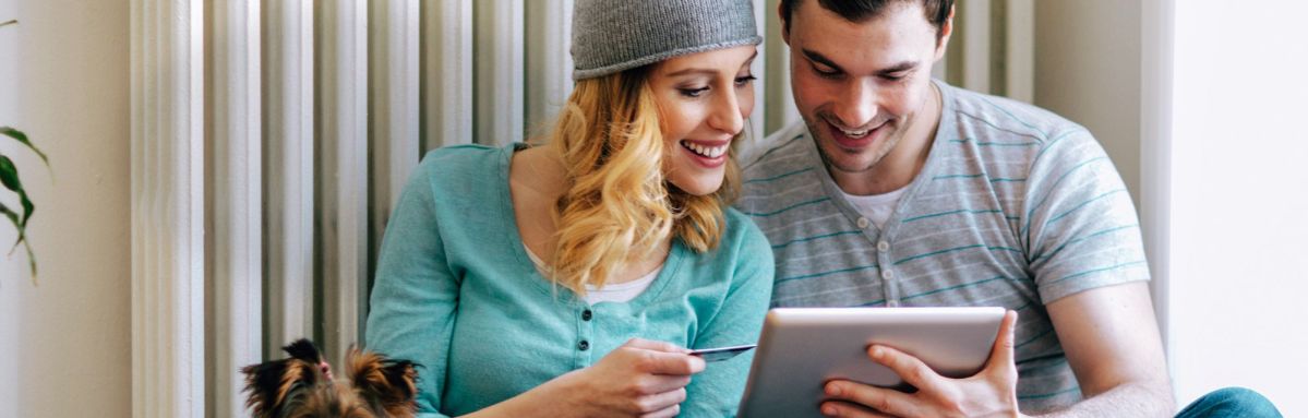 Eine Frau und ein Mann betrachten lächelnd ein Tablet zusammen, während die Frau eine Kreditkarte hält. Sie sitzen in einem Raum mit metallischen Wandpaneelen.