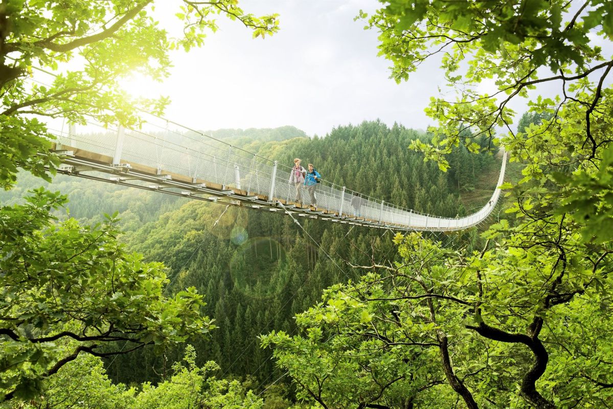 Eine Hängebrücke überquert ein bewaldetes Tal; zwei Personen spazieren darauf, umgeben von grünen Bäumen und sonnigem Himmel.