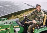 Ein lachender Mann fährt einen grünen Traktor vor großen, schrägen Solarpanelen auf einer Wiese. Er trägt einen gestreiften Pullover und eine Mütze.