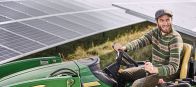 Ein lachender Mann fährt einen grünen Traktor vor großen, schrägen Solarpanelen auf einer Wiese. Er trägt einen gestreiften Pullover und eine Mütze.