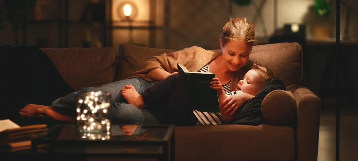 Eine Frau liegt mit ihrer Tochter auf einem Sofa und liest aus einem Buch vor. Auf dem Sofatisch steht ein Glas mit einer Lichterkette.