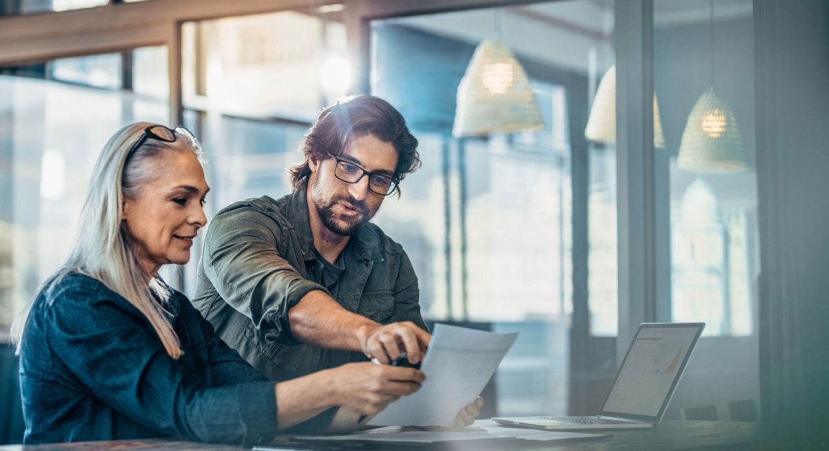 Eine Frau und ein Mann betrachten ein Dokument an einem Schreibtisch. Neben ihnen steht ein Laptop. Sie befinden sich in einem modernen, gut beleuchteten Büro mit großen Fenstern.