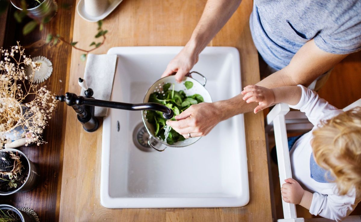 Familie wäscht Salat Eine Person wäscht frischen Spinat in einem Sieb unter dem Wasserhahn, während ein Kind zuschaut. Die Szene spielt sich in einer Küche mit Holztisch ab.
