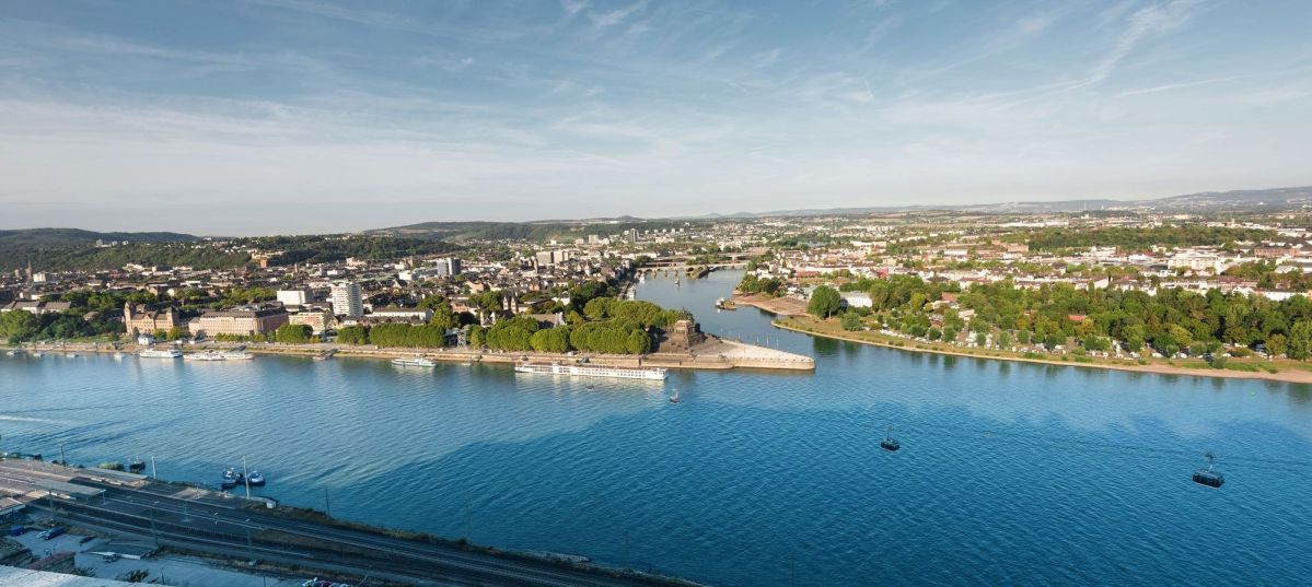 Deutsches Eck Koblenz Deutsches Eck Koblenz. Ein breiter Fluss fließt ruhig, gesäumt von grünen Bäumen und städtischen Gebäuden auf beiden Seiten, unter einem klaren blauen Himmel in einer weitläufigen Stadtlandschaft.