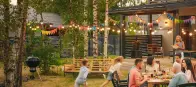 Eine Familie sitzt an einem Tisch und isst im Garten. Kinder rennen spielend herum. Bunte Lichterketten und Wimpel schmücken den Bereich zwischen Bäumen und einem modernen Haus.