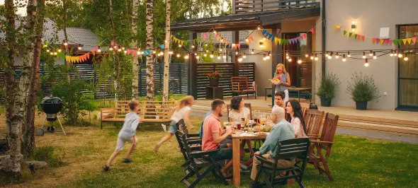 Eine Familie sitzt an einem Tisch und isst im Garten. Kinder rennen spielend herum. Bunte Lichterketten und Wimpel schmücken den Bereich zwischen Bäumen und einem modernen Haus.