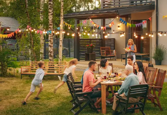 Eine Familie sitzt an einem Tisch und isst im Garten. Kinder rennen spielend herum. Bunte Lichterketten und Wimpel schmücken den Bereich zwischen Bäumen und einem modernen Haus.