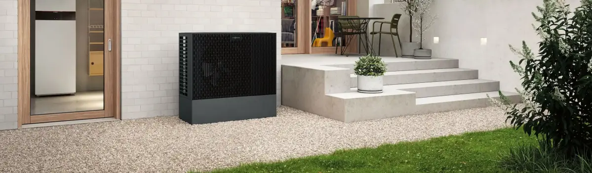 Eine dunkelgraue Wärmepumpe steht auf Kies, nahe einer Hauswand. Dahinter eine Terrasse mit Pflanzentopf, Tisch und Stühlen, vor einer Glastür im Hintergrund.