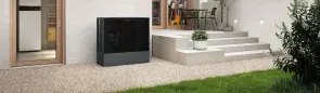 Eine dunkelgraue Wärmepumpe steht auf Kies, nahe einer Hauswand. Dahinter eine Terrasse mit Pflanzentopf, Tisch und Stühlen, vor einer Glastür im Hintergrund.