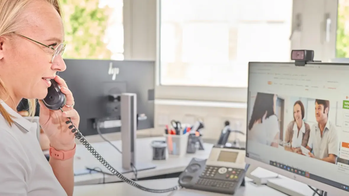 Eine Person telefoniert an einem Schreibtisch, während sie auf einen Monitor schaut, der eine evm-Webseite zeigt. Im Hintergrund befinden sich Fenster und Büroutensilien.