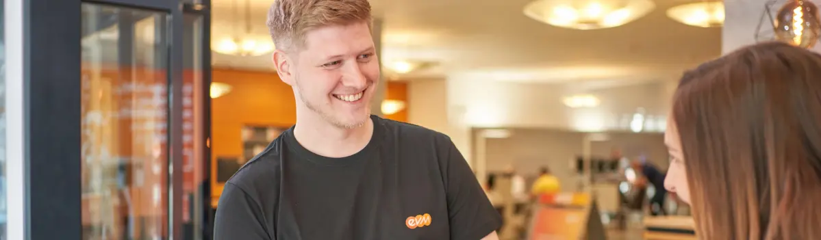 Ein lächelnder Mann trägt ein schwarzes T-Shirt mit orangefarbenem Logo ("evm"), steht in einem hell beleuchteten Raum mit anderen Personen und unscharfen Elementen im Hintergrund.